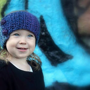May include: A young person wearing a purple and blue knitted headband with buttons. The headband is worn over their long brown hair. The person is smiling and looking at the camera. The background is a blue and green abstract design.