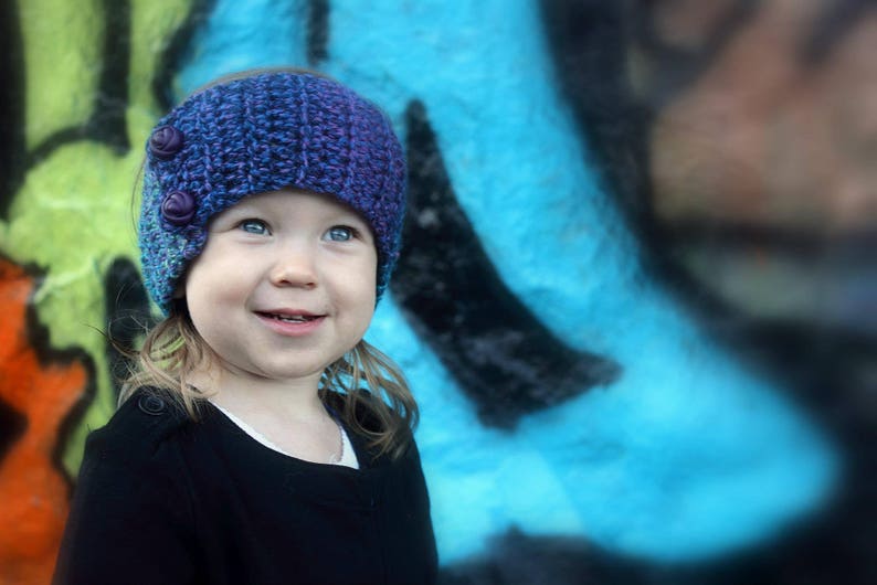May include: A young person wearing a purple and blue knitted headband with buttons. The headband is worn over their long brown hair. The person is smiling and looking at the camera. The background is a blue and green abstract design.