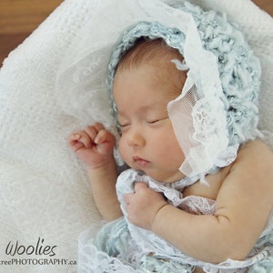 Op de afbeelding: Een slapende baby die een lichtblauwe gehaakte muts draagt met witte kant. De baby ligt op een wit deken in een rieten mand.