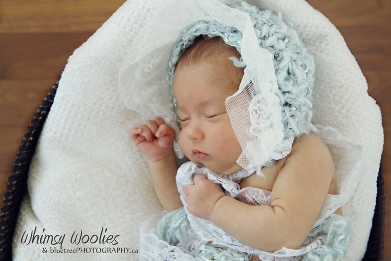 Op de afbeelding: Een slapende baby die een lichtblauwe gehaakte muts draagt met witte kant. De baby ligt op een wit deken in een rieten mand.