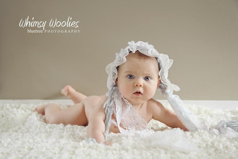 Op de afbeelding: Een baby met een witte kantmuts en een witte kantsjaal ligt op een wit, pluche oppervlak. De baby heeft blauwe ogen en kijkt naar de camera.