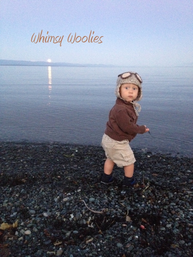 May include: A young child wearing a brown sweater and a brown and white aviator hat with goggles, standing on a rocky beach. The text "Whimsy Woolies" is written in orange at the top of the image.