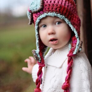 May include: A child wearing a pink, blue, and brown crocheted hat with long braids. The hat has a small, blue and brown square on top. The child is wearing a white sweater.