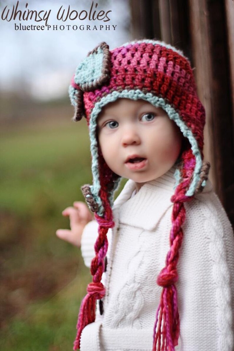 May include: A child wearing a pink, blue, and brown crocheted hat with long braids. The hat has a small, blue and brown square on top. The child is wearing a white sweater.