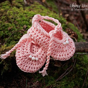 May include: A pair of pink crocheted baby shoes with pearl embellishments. The shoes are tied together with a pink crocheted ribbon.