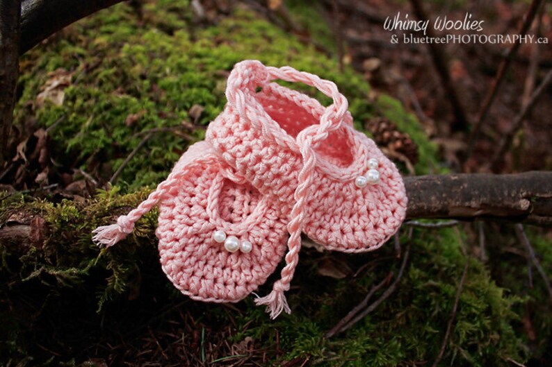 May include: A pair of pink crocheted baby shoes with pearl embellishments. The shoes are tied together with a pink crocheted ribbon.