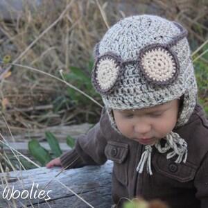 May include: A baby wearing a brown and beige crocheted aviator hat with a brown and beige crocheted goggles design. The hat has ear flaps and a long tassel.