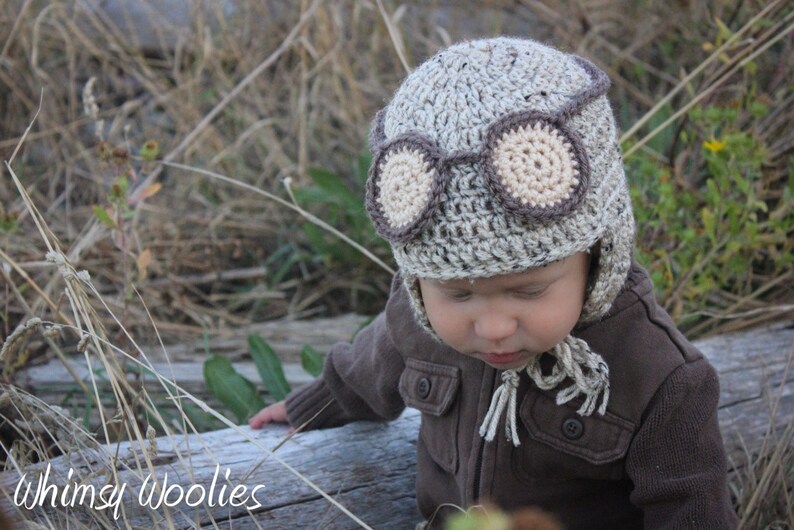May include: A baby wearing a brown and beige crocheted aviator hat with a brown and beige crocheted goggles design. The hat has ear flaps and a long tassel.