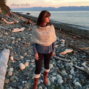 May include: A woman wearing a white and black knitted poncho, a blue denim shirt, black leggings, and white and brown leg warmers. She is standing on a rocky beach with a body of water and mountains in the background. The text "Whimsy Woolies" is visible in the top left corner of the image.