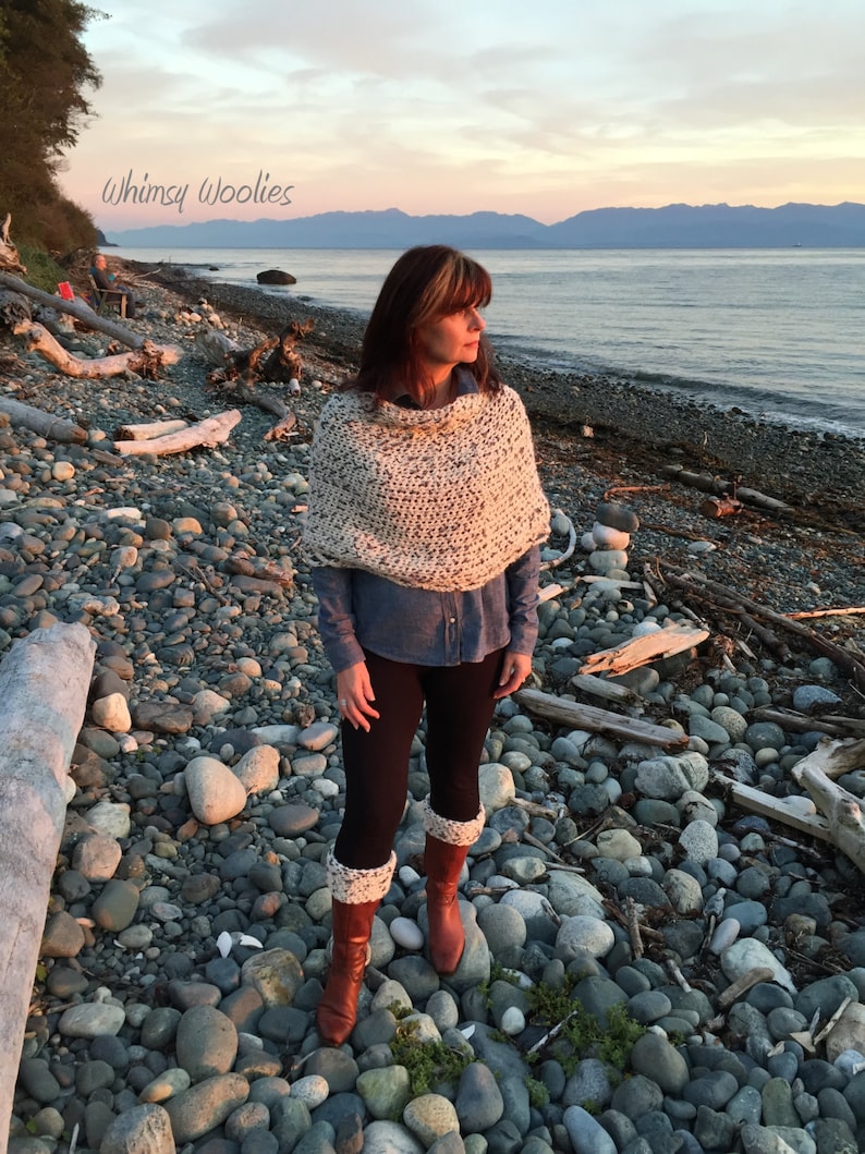 May include: A woman wearing a white and black knitted poncho, a blue denim shirt, black leggings, and white and brown leg warmers. She is standing on a rocky beach with a body of water and mountains in the background. The text "Whimsy Woolies" is visible in the top left corner of the image.
