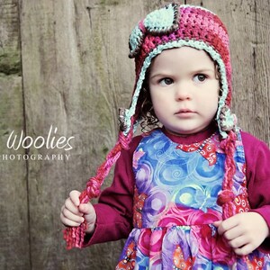 May include: A young child wearing a pink and blue crocheted aviator hat with a long tassel. The hat has a brown leather-like detail on the front. The child is wearing a colorful patterned dress and a maroon long-sleeved shirt. The background is a wooden wall with the text "Whimsy Woolies bluetree' PHOTOGRAPHY" written on it.