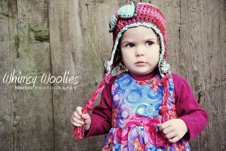 May include: A young child wearing a pink and blue crocheted aviator hat with a long tassel. The hat has a brown leather-like detail on the front. The child is wearing a colorful patterned dress and a maroon long-sleeved shirt. The background is a wooden wall with the text "Whimsy Woolies bluetree' PHOTOGRAPHY" written on it.