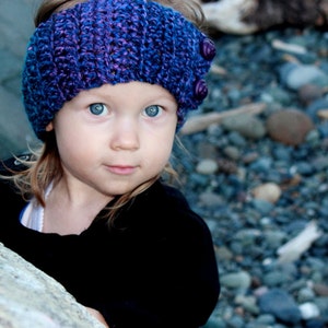 May include: A young girl wearing a purple and blue crocheted headband with buttons. The headband is a winter accessory for warmth and style.