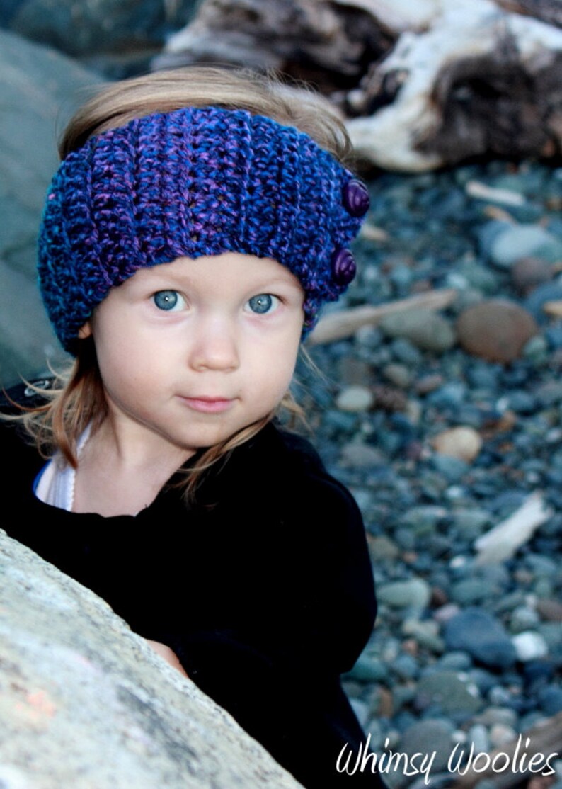 May include: A young girl wearing a purple and blue crocheted headband with buttons. The headband is a winter accessory for warmth and style.