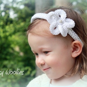 May include: A white crocheted headband with a flower design and a pearl accent. The headband is worn by a young child with blonde hair.