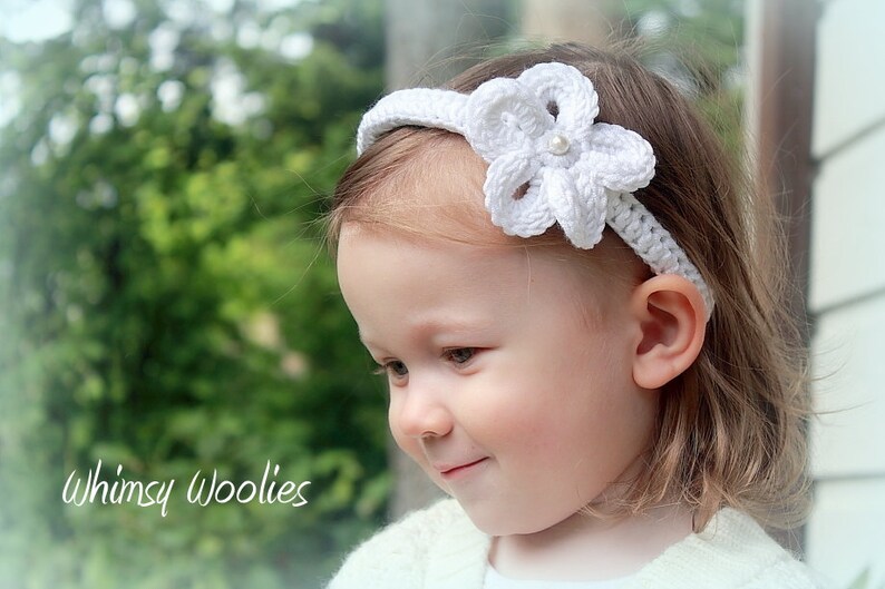 May include: A white crocheted headband with a flower design and a pearl accent. The headband is worn by a young child with blonde hair.