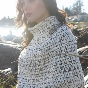 May include: A white and grey crochet poncho with a high neck and a fringed edge. The poncho is being worn by a person with long brown hair. The background is a blurred image of a beach.
