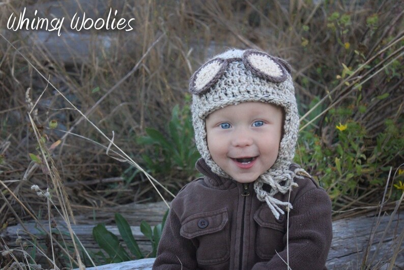 May include: A baby wearing a brown and beige crocheted aviator hat with goggles. The hat has a long scarf attached. The baby is smiling and looking at the camera.