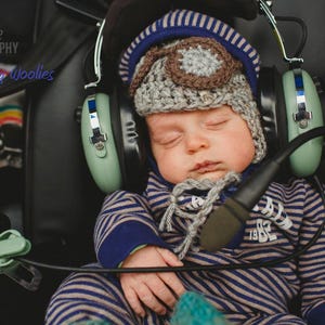 May include: A baby wearing a blue and brown knitted hat with earmuffs, sleeping in a stroller. The earmuffs are green and have a microphone attached.