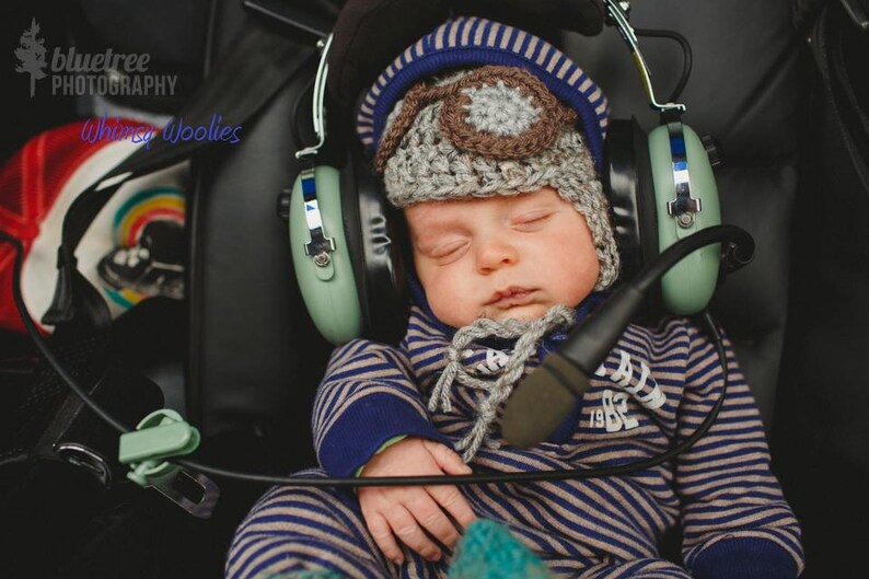 May include: A baby wearing a blue and brown knitted hat with earmuffs, sleeping in a stroller. The earmuffs are green and have a microphone attached.