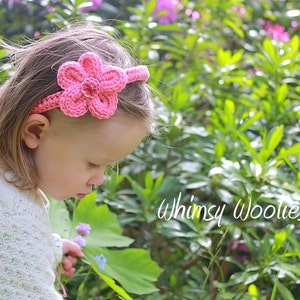 May include: A young girl wearing a pink crocheted headband with a flower design. The headband is on her forehead and the flower is on the side of her head. She is looking down at a flower in her hand. The background is a blurry image of green leaves and pink flowers.