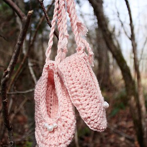 May include: A pair of pink crocheted baby booties with white pearl embellishments hanging from a tree branch.