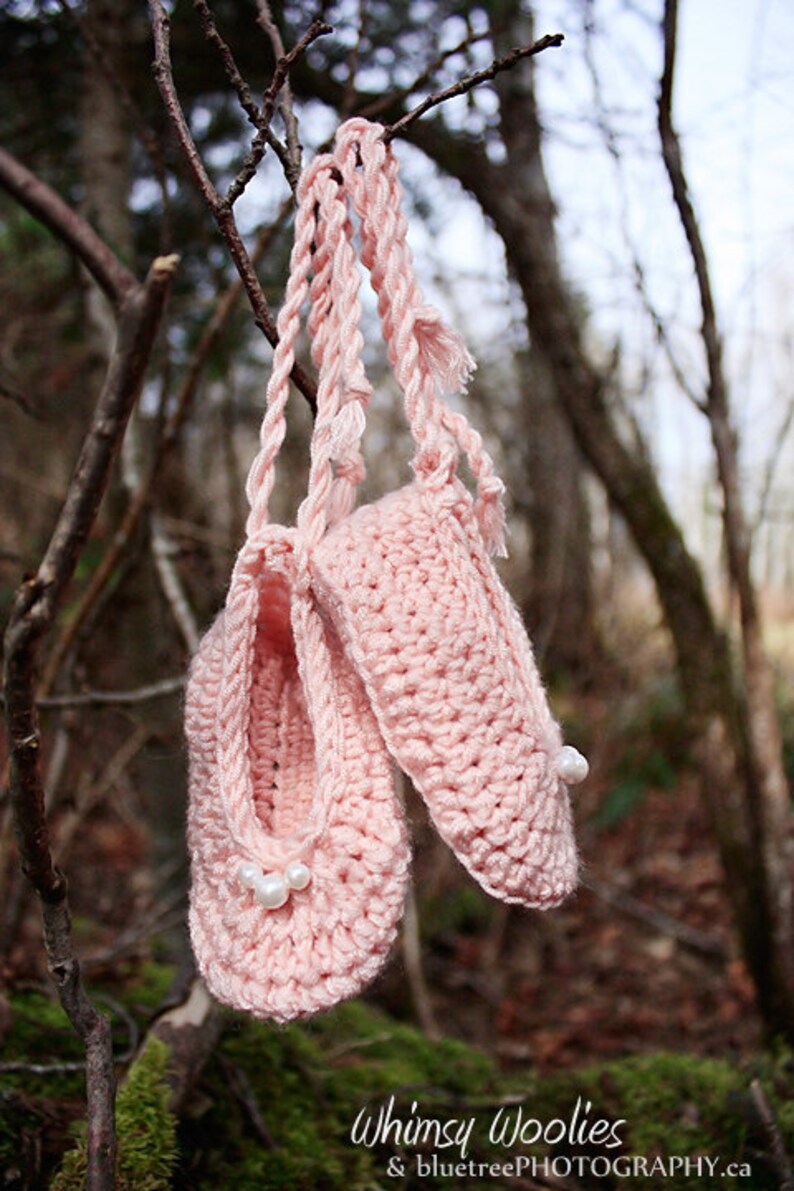 May include: A pair of pink crocheted baby booties with white pearl embellishments hanging from a tree branch.