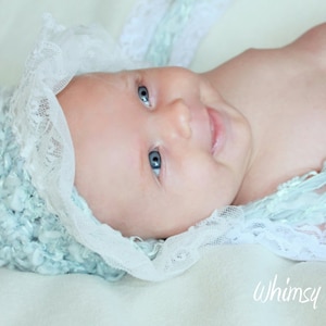 Op de afbeelding: Een baby die een lichtblauwe en witte gehaakte muts draagt met een kantversiering. De baby heeft blauwe ogen en lacht.