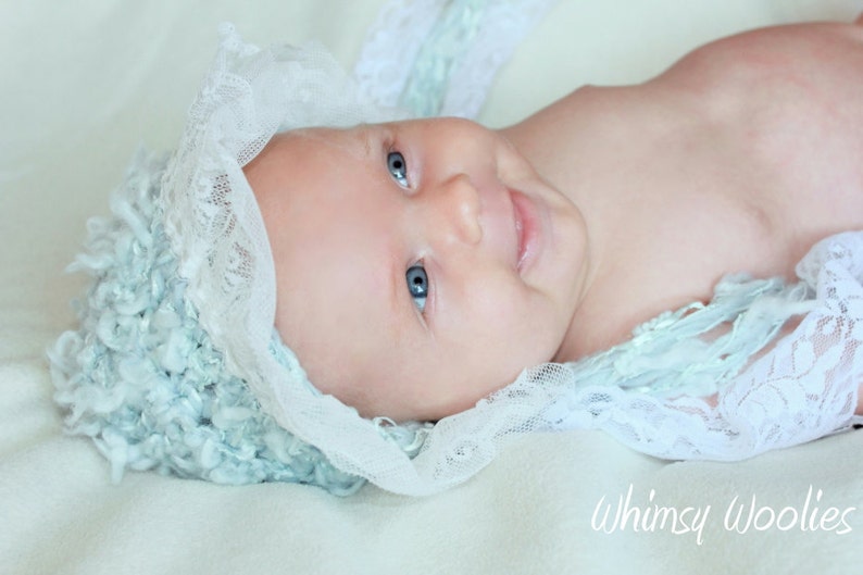 Op de afbeelding: Een baby die een lichtblauwe en witte gehaakte muts draagt met een kantversiering. De baby heeft blauwe ogen en lacht.