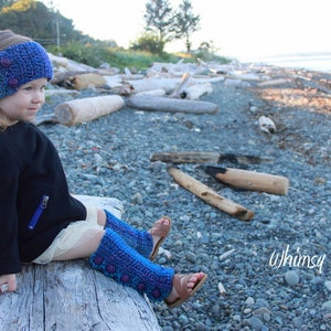 May include: A young person wearing a blue and purple crocheted leg warmer and a blue and purple crocheted headband with buttons. The person is sitting on a log on a beach with driftwood and rocks in the background. The text "Whimsy Woolies" is visible in the lower right corner of the image.