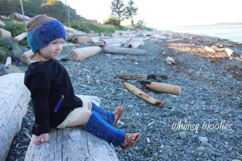 May include: A young person wearing a blue and purple crocheted leg warmer and a blue and purple crocheted headband with buttons. The person is sitting on a log on a beach with driftwood and rocks in the background. The text "Whimsy Woolies" is visible in the lower right corner of the image.