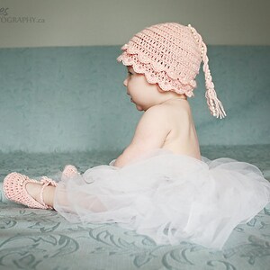 May include: A baby wearing a pink crocheted hat with a long tail and a white tulle skirt sits on a blue patterned bed.