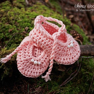May include: A pair of handmade, light pink crochet baby shoes with pearl accents. The shoes are tied together with a braided cord and are displayed on a mossy branch. The image includes the text "Whimsy Woolies & bluetreePHOTOGRAPHY.ca".