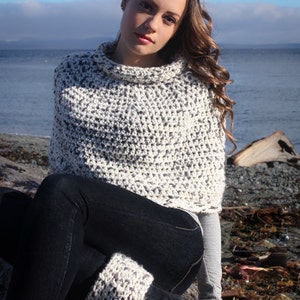 May include: A woman wearing a white and grey crocheted poncho with a turtleneck collar. She is sitting on a log with her legs crossed and wearing black jeans and grey knitted leg warmers. The background is a beach with a blue sky and water.