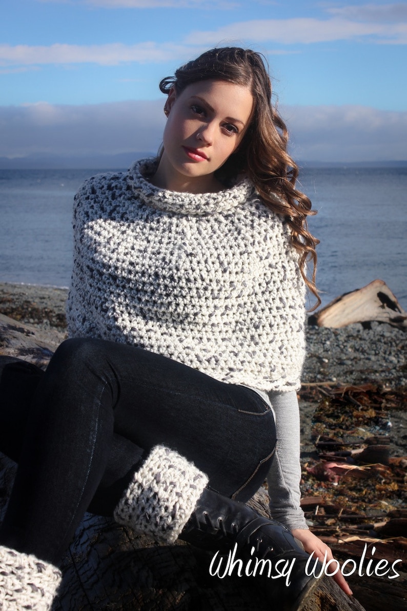 May include: A woman wearing a white and grey crocheted poncho with a turtleneck collar. She is sitting on a log with her legs crossed and wearing black jeans and grey knitted leg warmers. The background is a beach with a blue sky and water.