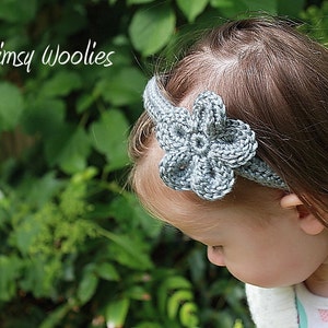 May include: A gray crocheted headband with a flower design. The headband is worn by a child with long brown hair. The background is a blurred image of green foliage.
