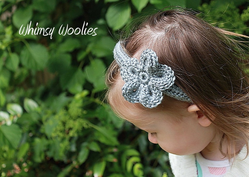 May include: A gray crocheted headband with a flower design. The headband is worn by a child with long brown hair. The background is a blurred image of green foliage.
