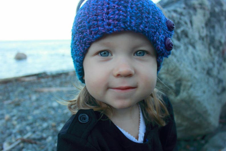 May include: A young person wearing a purple and blue knitted hat with a flower design. The hat has two large purple buttons on the side. The person is wearing a black jacket.