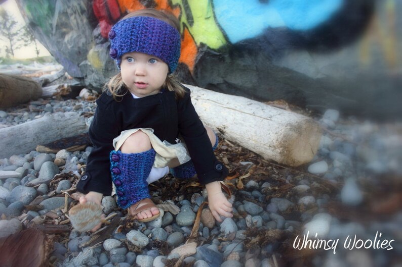 May include: A young girl wearing a purple and blue crocheted hat with a black jacket and matching leg warmers. The girl is sitting on a rock with a blurred background of a colorful wall.