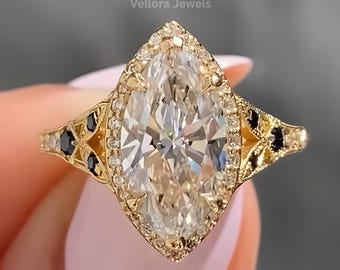 May include: A marquise-cut diamond ring with a gold band. The ring has a halo of smaller diamonds around the central stone and black accent stones. The ring is held up by a person with pink nail polish.