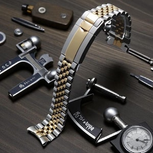 May include: A two-tone stainless steel and gold watch band with a jubilee-style link design. The band is displayed with watch repair tools and accessories on a dark wood surface. The band features a gold center link and a silver outer link.