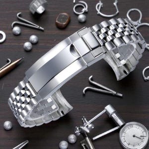 May include: A stainless steel watch band with a polished finish. The band is composed of small, rectangular links and features a folding clasp. Various watchmaking tools and components are scattered around the band on a dark surface.