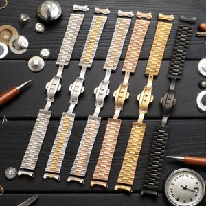 Puede incluir: Una colección de correas de reloj en varios acabados metálicos, incluyendo plata, oro, oro rosa y negro. Las correas se muestran sobre una superficie oscura, mostrando sus diferentes colores y estilos. Herramientas y componentes de relojería están dispersos.
