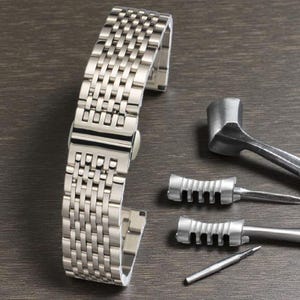May include: A silver stainless steel watch band with a woven design. The band is next to watch repair tools, including a hammer and several pin removal tools. The items are on a dark wood surface.