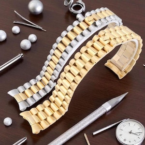 May include: Two-tone watch straps in gold and silver, presented on a dark wood surface. The straps are curved, highlighting their design. Various watch repair tools and small decorative spheres surround the straps, suggesting a watch repair or accessory theme.
