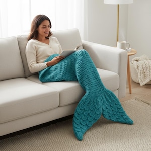 May include: A teal mermaid tail blanket with a textured, scale-like design. The blanket is made of a soft, knitted material and is designed to be worn while relaxing. The tail features a fan-shaped fin. The blanket is shown on a white sofa.