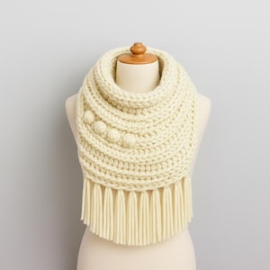 May include: Cream-colored, chunky knit cowl scarf with three decorative bobbles on the side. The scarf has a fringed bottom edge and is displayed on a mannequin. The thick knit suggests warmth and a cozy feel.