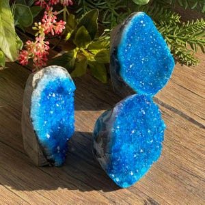 May include: Three blue geode crystals with a sparkling, crystalline interior. The geodes have a rough, gray exterior and are arranged on a wooden surface. The crystals are a vibrant shade of blue, with a natural, organic shape.