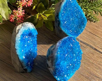 May include: Three blue geode crystals with a sparkling, crystalline interior. The geodes have a rough, gray exterior and are arranged on a wooden surface. The crystals are a vibrant shade of blue, with a natural, organic shape.