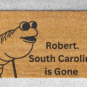 May include: A rectangular doormat with a cartoon frog illustration and the text "Robert. South Carolina is Gone" in black. The doormat is made of a light brown material.
